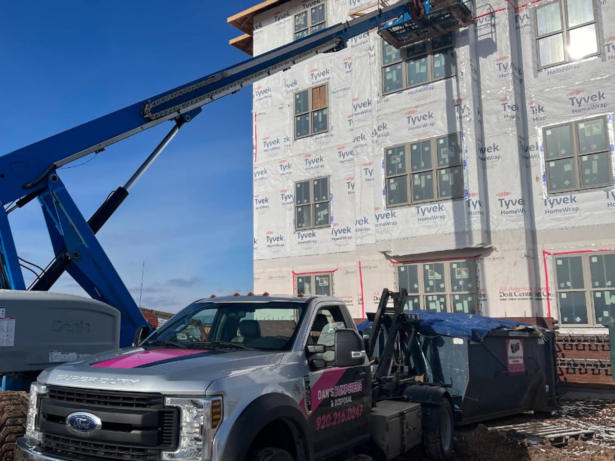 A Dan's Dumpsters hooklift truck placing a roll-off dumpster beside a multi-story building under construction