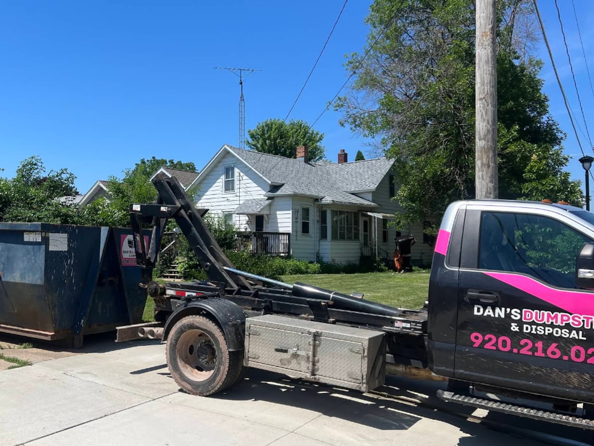 A pink-lettered Dan's Dumpsters truck delivering a roll-off dumpster in a residential neighborhood